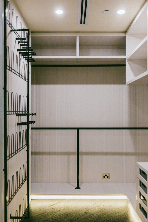 A compact custom walk-in wardrobe with white built-in shelving, upper storage compartments, hanging rails, and a decorative black metal-framed door or screen. Integrated warm LED strip lighting illuminates the lower shelving and pale timber-look floor, while recessed ceiling lights provide even illumination. The scheme is minimalist with contrasting black hardware and linear detailing.