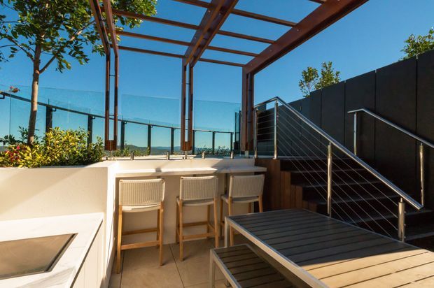 Contemporary rooftop or elevated outdoor entertaining area with a built-in white bar counter and three stools, timber-framed glass pergola, glass balustrades, and a stairway with stainless-steel cable railing. A dark feature wall and timber dining table create a modern, refined outdoor setting with expansive sky views.