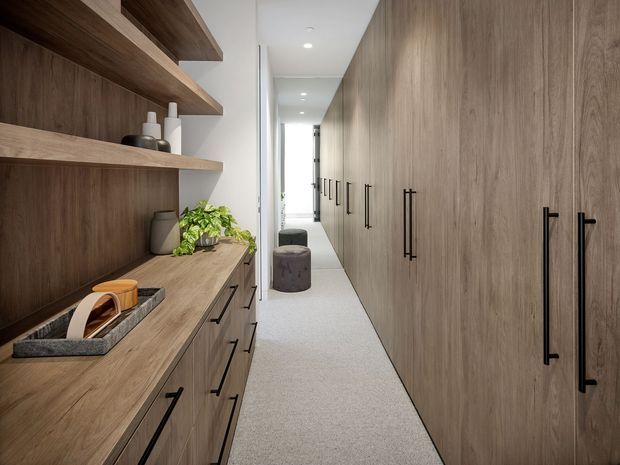 A narrow, contemporary walk-in wardrobe or dressing corridor with extensive dark timber-look built-in cabinetry on both sides. The left side features open wood shelves, drawers, a long countertop, decorative objects and a plant, while the right side has full-height wardrobes with long black handles. A full-height mirror extends down the corridor, reflecting the cabinetry and a small upholstered ottoman. Recessed ceiling lights and a pale carpet create a clean, refined, softly lit interior.
