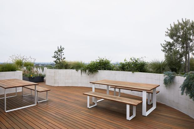 A contemporary rooftop terrace or elevated outdoor dining area with warm timber decking, two minimalist timber dining tables with integrated benches, and white rendered or masonry planter walls filled with grasses and shrubs. The open terrace overlooks a distant urban landscape beneath an overcast sky, creating a clean, relaxed outdoor setting.