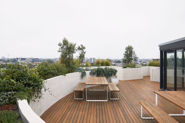 A contemporary rooftop terrace with extensive timber decking, built-in white rendered planter walls, lush planting, and a large outdoor dining table with bench seating. A glazed dark-framed pavilion or interior room opens onto the deck at right, with a low-rise urban landscape visible beyond.