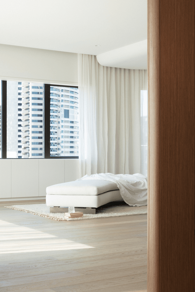 Bright minimalist bedroom with a low white upholstered bed, sheer floor-to-ceiling curtains, large dark-framed windows, pale timber flooring, and a sculptural curved ceiling detail. A high-rise urban view is visible outside, while a warm timber door or wall panel frames the right side of the image.