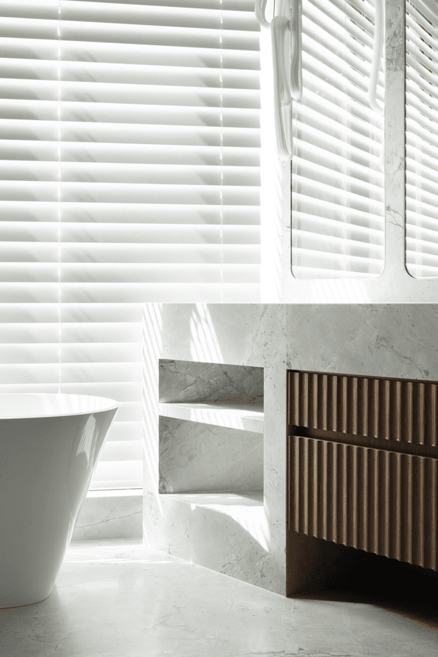 A bright, minimalist bathroom corner with a freestanding white bathtub, white marble-look wall and floor surfaces, open built-in shelving, and a dark fluted timber vanity or storage unit. White horizontal blinds create strong filtered sunlight and shadows across the space.