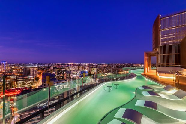 A rooftop pool area at dusk overlooking a brightly illuminated urban skyline. The pool has a curving layout with shallow lounging zones and sculptural in-water seating, bordered by transparent glass balustrades. A tall contemporary building structure with vertical screening and warm architectural lighting frames the right side.