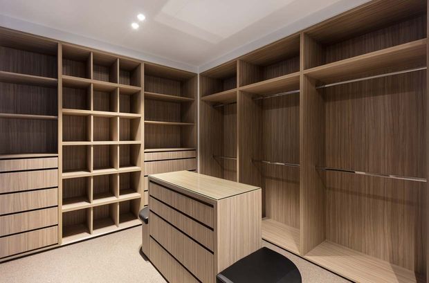 A spacious, custom-built walk-in wardrobe with extensive warm wood-grain cabinetry, open shelving, drawers, hanging rails, and a central island unit. Recessed ceiling lighting illuminates the fitted storage, with a small black upholstered stool in the foreground.