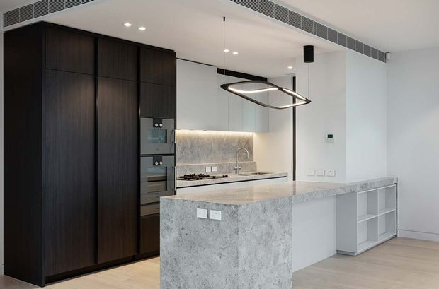 Contemporary open-plan kitchen with a large waterfall-edge stone island, dark full-height cabinetry, integrated appliances, pale stone benchtops and backsplash, and minimalist pendant lighting. A light timber-toned floor and white walls create a clean, refined interior.