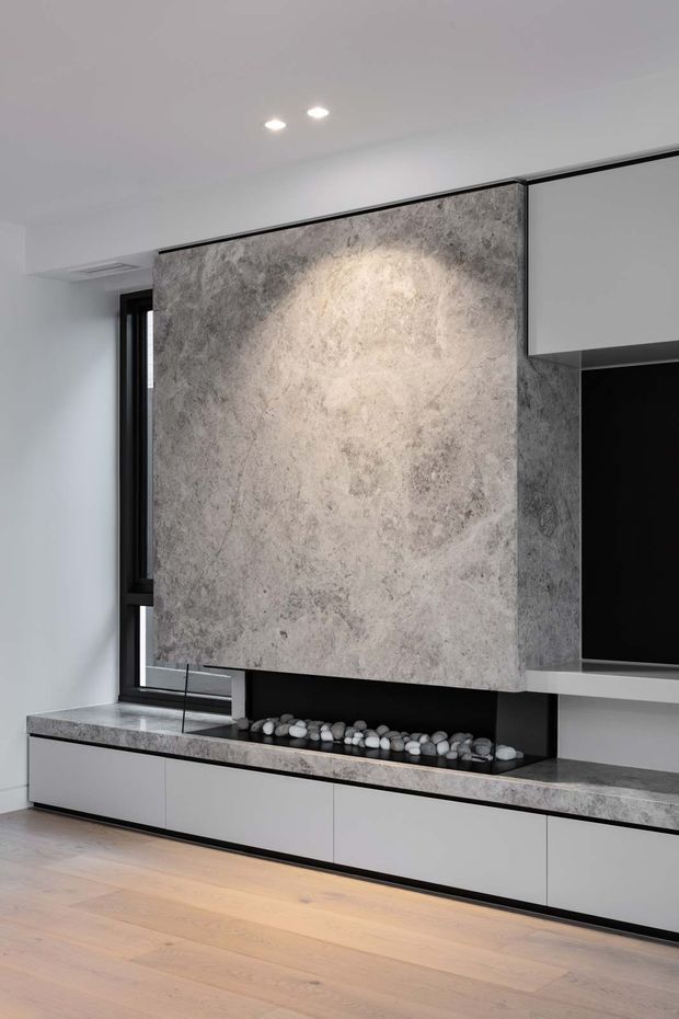 A contemporary built-in living room media wall with a large grey stone-look feature panel, black recessed niche, floating white cabinetry, and a matching stone benchtop. A linear decorative pebble bed sits beneath the panel, with black-framed glazing to the side and pale timber flooring.