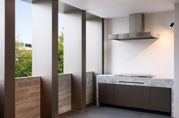 Minimalist contemporary kitchen with a dark built-in island or cabinet run, pale stone benchtop, integrated cooktop, and a large stainless-steel rangehood. Tall narrow windows between deep structural wall piers provide abundant natural light and views to greenery. The room uses restrained grey, white, and natural timber-toned finishes.