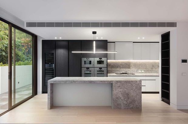 Contemporary minimalist kitchen with a large stone island, integrated dark cabinetry, pale upper cabinets, illuminated stone splashback, built-in appliances, and floor-to-ceiling glazing opening to greenery. A linear pendant light and concealed ventilation grille reinforce the clean, refined design.
