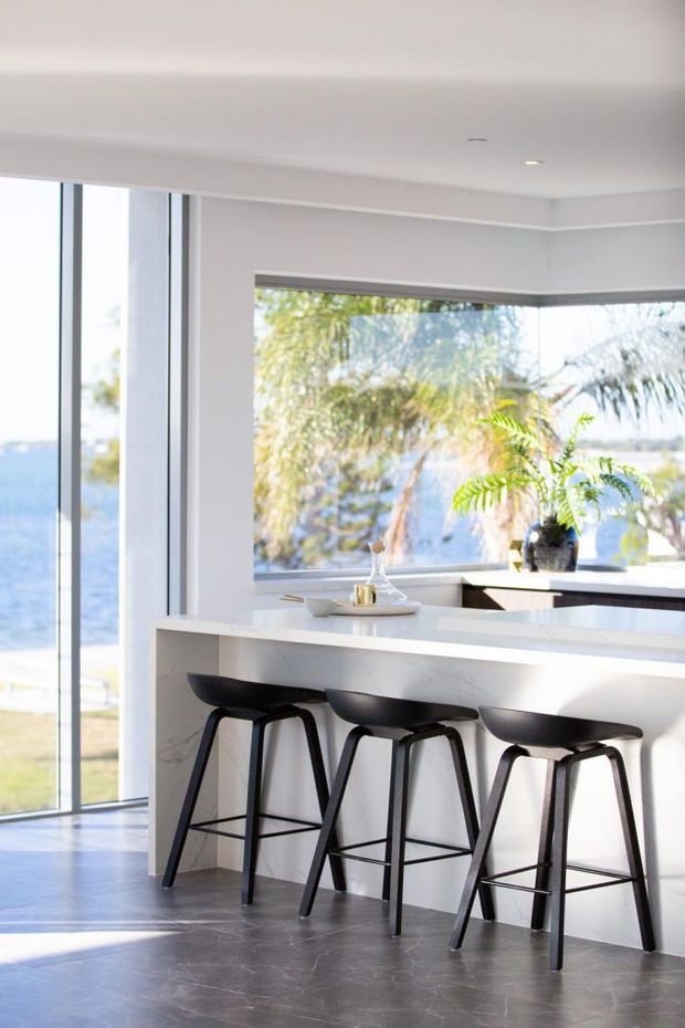 Bright contemporary kitchen with a white island and waterfall-style benchtop, three black bar stools, extensive floor-to-ceiling glazing, and a large corner window overlooking water and greenery. The space has a clean, airy feel with dark stone-look flooring and minimal recessed lighting.