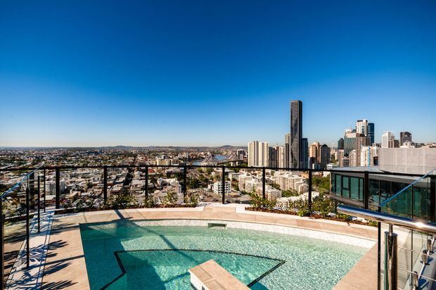 Rooftop pool terrace overlooking a dense urban skyline. The image shows a turquoise swimming pool with a shallow sun shelf and central platform, pale paving, glass balustrades, lounge seating, and a modern city apartment tower backdrop under a clear blue sky.