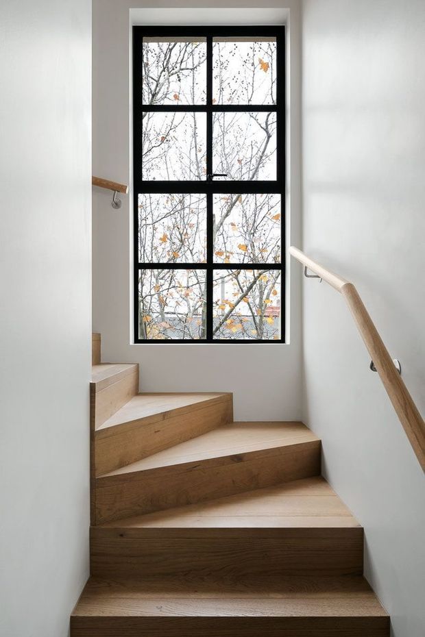 A narrow interior stairway with warm timber-clad treads and risers ascending toward a tall black-framed multi-pane window. White walls and simple timber handrails create a clean, bright, understated composition, with bare tree branches visible outside.