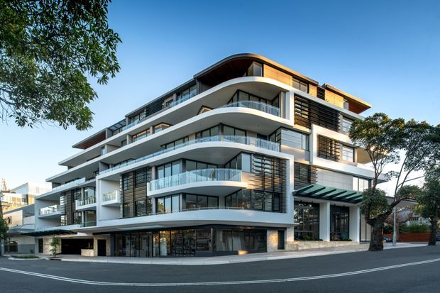 Contemporary multi-storey apartment building viewed from the street, with layered curved balconies, extensive floor-to-ceiling glazing, dark horizontal screening, white rendered forms, and warm timber-toned soffits and upper-level cladding. The building occupies a landscaped urban corner site with a recessed ground-level entry and parking access.