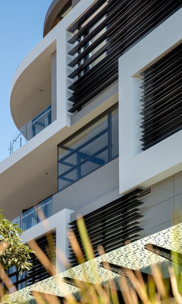 Vertical exterior architectural photograph of a contemporary multi-storey building with white rendered walls, curved projecting balconies, blue-tinted glazing with dark frames, and extensive black horizontal louvre screens used as sun shading. A patterned privacy or shade screen and planted foreground are visible at the base, with clear blue sky behind.