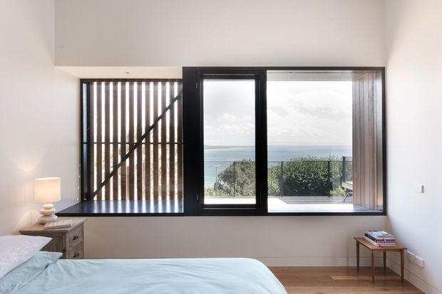 A minimalist coastal bedroom viewed from inside, with a low bed, pale bedding, timber flooring, a small bedside table and lamp, and a broad black-framed window assembly. Vertical timber screening and glazing frame views toward the ocean and landscaped coastal vegetation.