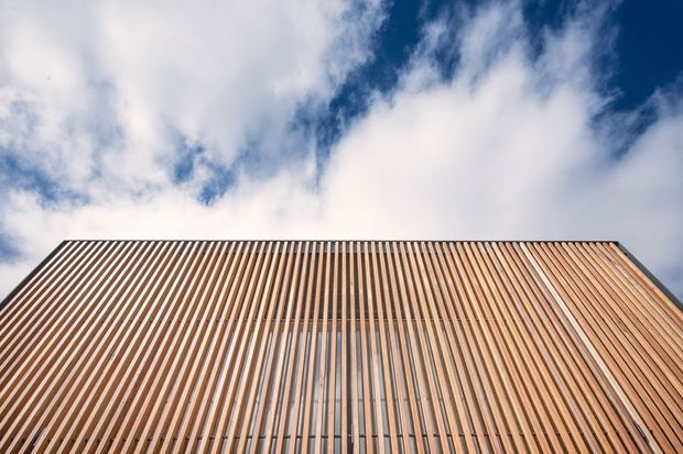 Upward-looking architectural detail of a building facade clad in closely spaced vertical timber battens, creating a strong linear rhythm against a partly cloudy blue sky. The image emphasizes the facade geometry and warm wood tones.