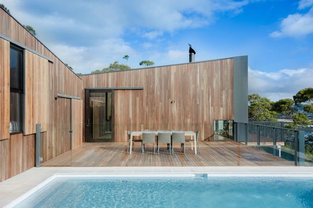 Contemporary timber-clad residence with a broad timber deck overlooking a swimming pool. The image focuses on an outdoor dining terrace enclosed by frameless glass balustrades, with dark-framed glazing and a simple flat-roofed architectural form. Blue sky, mature trees, and a distant coastal-looking outlook are visible beyond the property.