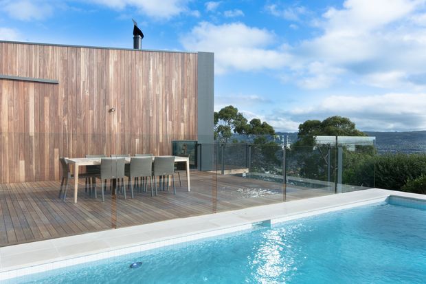 Exterior view of a contemporary pool terrace overlooking a coastal or waterfront landscape. A large turquoise swimming pool occupies the foreground, bordered by pale paving and a timber deck with an outdoor dining table and chairs. The building has a simple flat-roofed form with vertical timber cladding and a dark metal flue, while frameless glass balustrades provide open views beyond.