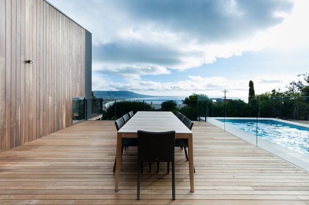 Contemporary outdoor dining area on a timber deck beside a swimming pool, with a long dining table and dark chairs, vertical timber-clad wall, frameless glass balustrades, and views toward the water and distant hills.
