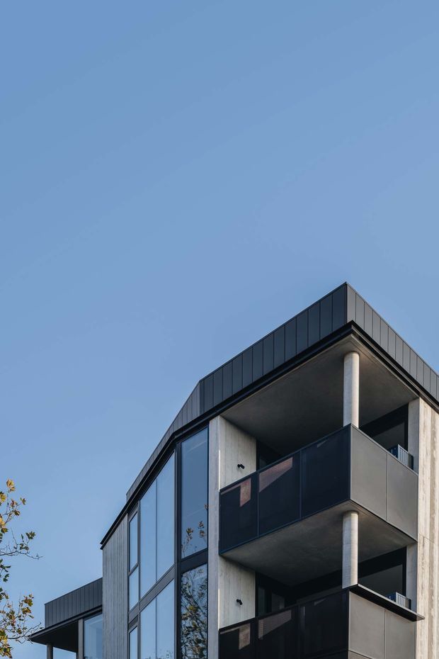 Low-angle exterior view of a contemporary multi-storey residential building with a dark standing-seam roof edge, large vertical glazed openings, raw concrete walls and columns, and stacked balconies enclosed by dark privacy screens. The building is set against a clear blue sky with minimal surrounding vegetation visible.