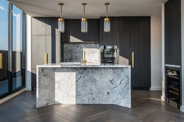 Contemporary kitchen featuring a large marble-look island with waterfall ends, dark timber cabinetry, integrated appliances, brass pendant lights and hardware, and dark herringbone timber flooring. Full-height glazing brings in abundant natural light.