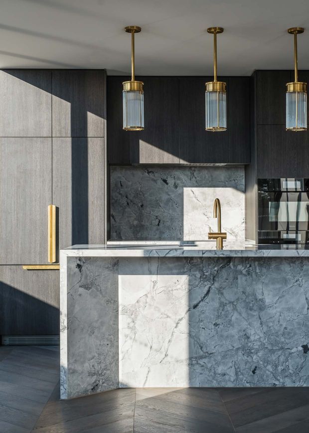 A sophisticated contemporary kitchen featuring dark timber cabinetry, a substantial grey-veined stone island and matching splashback, integrated appliances, and a brass-toned faucet. Three fluted glass pendant lights with brass detailing hang above the island. Strong natural light creates geometric highlights across the cabinetry, stone surfaces, and dark herringbone floor.
