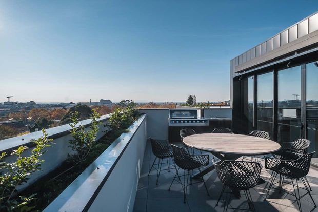 A contemporary rooftop terrace with a round outdoor dining table and wire-frame chairs, bordered by raised planter beds. The terrace connects to a dark-framed glazed interior and includes a built-in outdoor cooking area, with expansive urban views beyond.