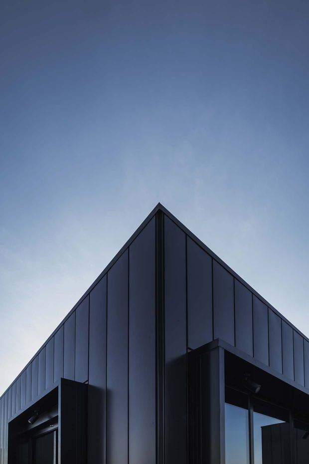 Low-angle exterior architectural photograph of a sharply peaked, dark-clad building corner. Vertical metal facade panels, deep dark window and door openings, and strong geometric roof and wall lines create a dramatic minimalist composition against a blue evening sky.