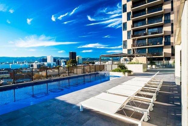 A sunny rooftop or elevated pool terrace beside a modern multi-storey apartment building. The image shows a long blue swimming pool with dark tiled surrounds, rows of white sun loungers, metal balustrades, planted raised beds, and a city skyline with hills in the distance. The building has a contemporary rectilinear facade with balconies and extensive glazing.
