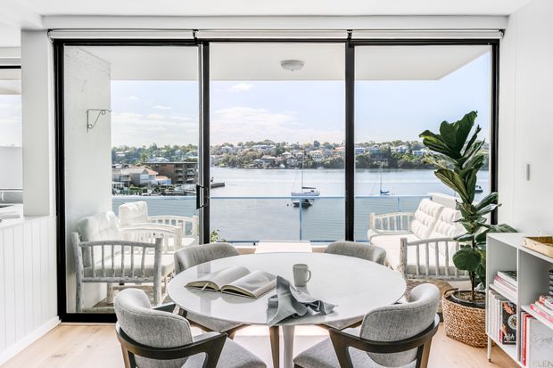 Bright contemporary dining area with a round dining table and upholstered chairs positioned beside full-height black-framed glazing. The room opens to a waterfront view with boats and houses across the water. White walls, light timber flooring, built-in shelving, and a large potted plant create an airy, refined interior.
