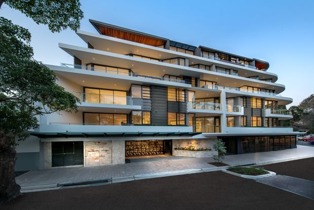 Exterior architectural photograph of a contemporary multi-storey apartment building with layered cantilevered balconies, extensive glazing, horizontal bands, dark louvres, warm timber soffits, and a rooftop terrace. The building fronts a paved driveway and landscaped street edge, with ground-level parking or garage openings visible.