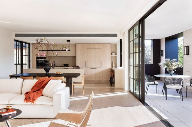 Bright open-plan interior combining a living room, dining area and kitchen, opening through large black-framed glazed doors to a covered outdoor dining terrace. The space features pale timber built-in kitchen cabinetry, light flooring, white walls, a white sofa with a rust-orange throw, a dark dining table, and abundant natural light. The design is clean-lined and contemporary with strong indoor-outdoor connection.