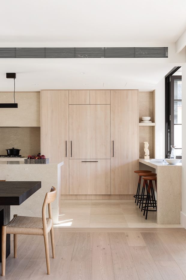 Bright, refined contemporary kitchen and dining area with pale timber flooring and full-height light timber cabinetry. A stone-look kitchen run includes a cooktop at left, while a peninsula or island with dark benchtop occupies the foreground. A pale stone breakfast bar with several dark-framed timber stools sits beside black-framed glazing. The room has a clean white ceiling with a long linear ventilation grille and minimalist pendant lighting.