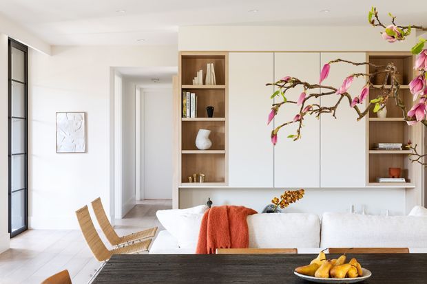 Bright open-plan living and dining area with a white upholstered sofa, dark timber dining table, woven timber dining chairs, and a full-height built-in timber and white cabinetry wall. Large black-framed glazing is visible at left, while sculptural artwork, books, ceramics, and flowering branches add decorative detail. The space has a clean, refined contemporary appearance with abundant natural light.
