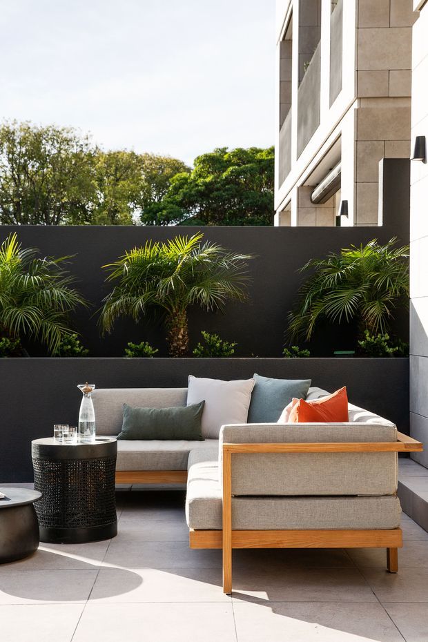 A bright, contemporary outdoor lounge courtyard beside a modern building. The space features a large L-shaped upholstered sofa with timber framing, a black perforated cylindrical side table, pale large-format tiled paving, dark rendered planter walls, and layered tropical planting. The building facade has light stone or concrete panels, deep reveals, and modern wall lighting. Strong sunlight creates crisp shadows, giving the restrained neutral palette a warm, inviting character with orange and green accents.