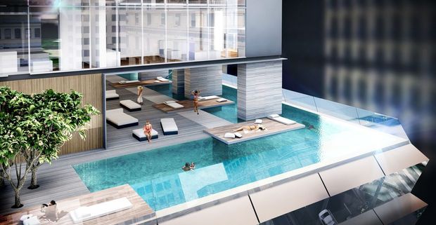 Architectural rendering of a rooftop swimming pool and resort-style outdoor amenity area atop a contemporary multi-storey building. The scene includes a large turquoise pool with stepped and shallow lounging zones, timber-look decking, sun loungers, integrated cabanas or seating bays, privacy screens, planted trees, and surrounding urban apartment or office façades. The image is presented from an elevated exterior viewpoint with strong geometric forms and a clean modern aesthetic.