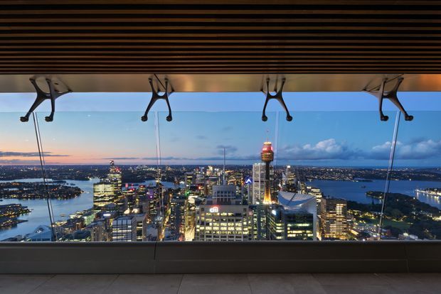 Panoramic elevated view of a brightly lit city skyline and surrounding harbour at dusk, seen through expansive glazing or an open viewing platform. A dark horizontal slatted ceiling and suspended structural or cable elements frame the upper portion, with a simple dark sill and tiled floor in the foreground.