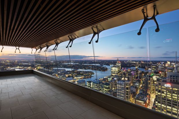 A high-level covered terrace or balcony with expansive floor-to-ceiling glazing and panoramic views over a brightly lit urban waterfront at dusk. The space has large-format grey stone or porcelain floor tiles, a dark linear slatted ceiling, and distinctive exposed curved metal support brackets along the glazed edge. The scene feels elevated, contemporary, and dramatic.