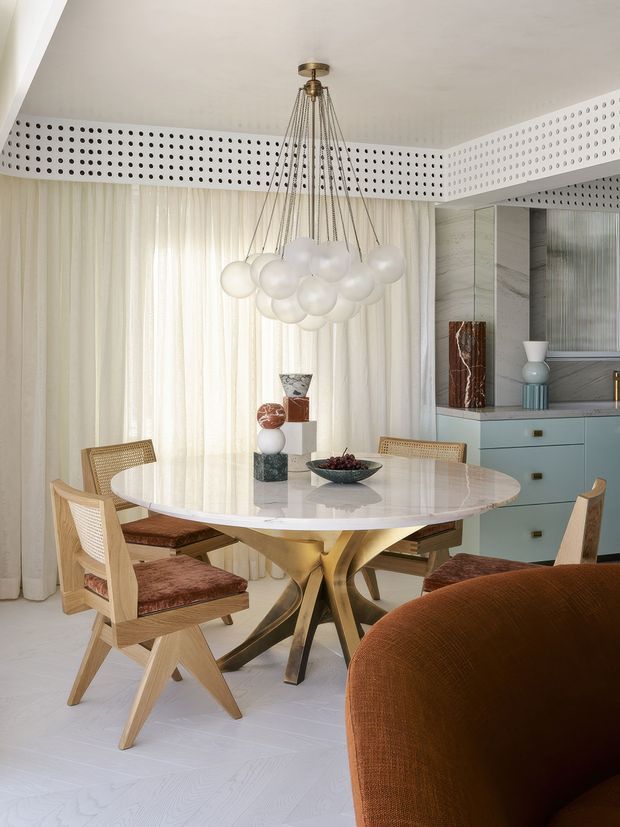 A refined dining area with a round marble-topped table and sculptural metallic pedestal base, surrounded by mid-century-inspired wooden dining chairs with woven backs and rust-coloured upholstery. A dramatic clustered globe pendant hangs above, while sheer floor-to-ceiling curtains, a perforated decorative ceiling band, and pale blue built-in cabinetry create a bright, elegant interior. Part of a curved rust-coloured sofa is visible in the foreground.