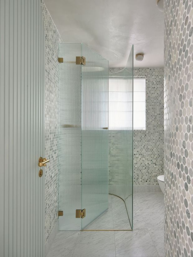 A narrow contemporary bathroom featuring a frameless fluted-glass shower enclosure with brass hardware, pale grey and white pebble-pattern mosaic wall tiles, vertically grooved wall panelling, and large light marble-look floor tiles. A toilet is partially visible beyond the shower, with bright filtered daylight from the window.