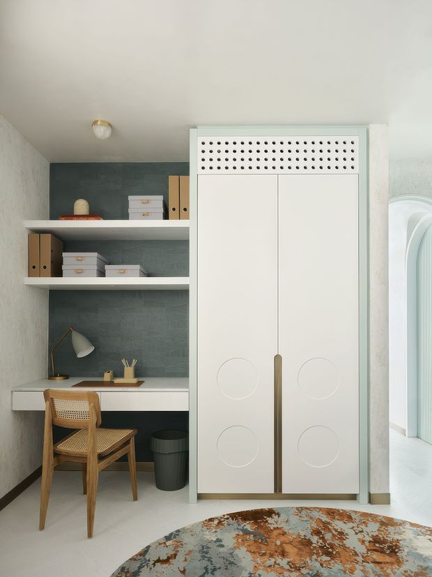 Compact, refined home office nook with a built-in white desk, open shelving, filing boxes, desk lamp and a woven wood-and-cane chair. The cabinetry features tall white doors with circular detailing, a slim brass-toned pull and perforated ventilation panel above. A dark blue-green textured wall backs the workspace, while an arched opening and pale textured walls add architectural character. A patterned round rug occupies the foreground.