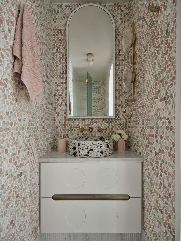 A compact, highly decorative bathroom vanity alcove clad in rounded pebble-pattern mosaic tiles. A white floating vanity with oversized brass handles supports a terrazzo vessel basin, brushed brass wall-mounted taps, and a tall arched mirror. Pink towels and matching accessories add a soft accent, while sculptural brass wall fixtures and floral decoration complete the refined, playful fit-out.