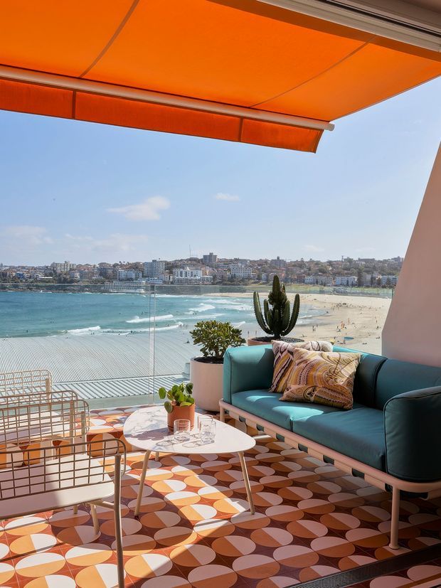 A bright, colourful seaside terrace or balcony overlooking a sandy beach, turquoise water, and a coastal urban shoreline. The space features a teal outdoor sofa, pale pink metal lounge chairs, a small white coffee table, potted plants, patterned geometric floor tiles, and a large orange retractable awning. Full-height glazing or a glass balustrade provides an unobstructed coastal view. The interior styling is playful, retro-inspired, and highly saturated.