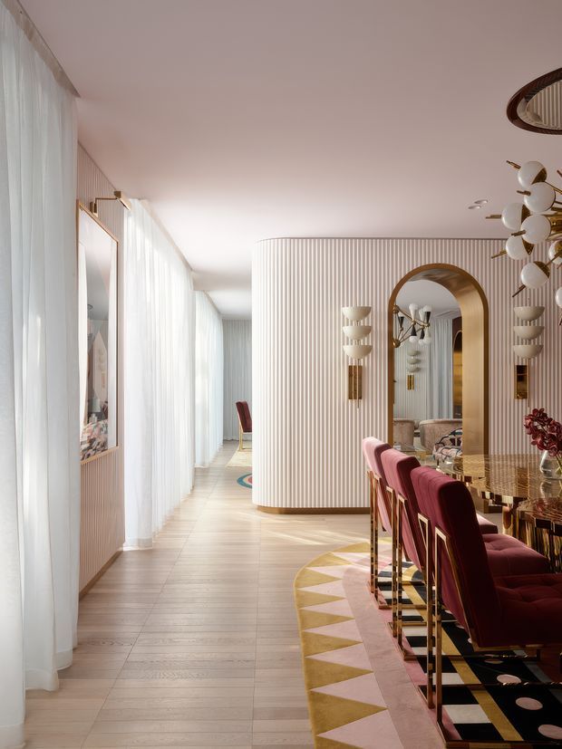 A sophisticated open-plan dining and living area with a long dining table, plush burgundy upholstered chairs, and a large arched mirror framed in warm metallic wood tones. The space features full-height sheer curtains, pale timber flooring, curved fluted wall panelling, sculptural globe lighting, brass accents, and a geometric patterned rug. The overall composition is glamorous, refined, and softly lit.