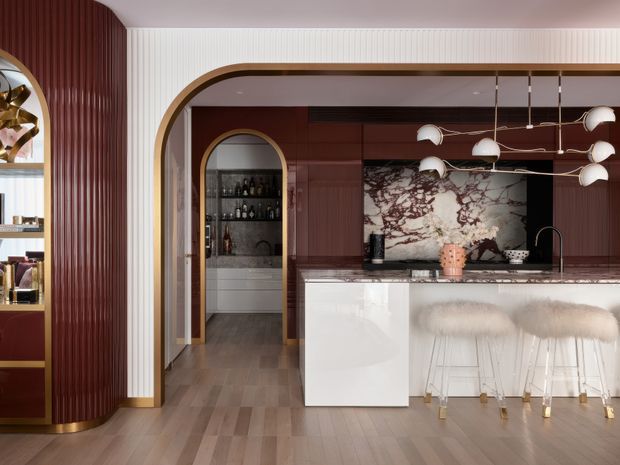 Refined contemporary kitchen and bar area with a dramatic veined stone splashback, glossy white island cabinetry, dark timber-toned cabinetry, fluted wall panelling, and a rounded arched opening framed in timber. A sculptural brass-and-white pendant light hangs above the island, which is paired with upholstered bar stools. The adjoining drinks bar and reflective finishes create a luxurious, polished interior.