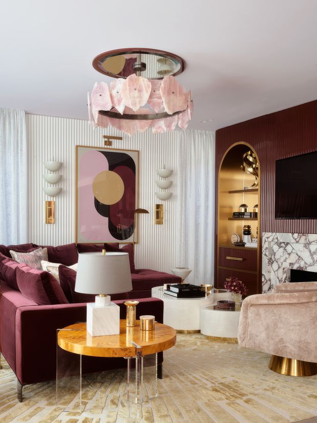 A richly styled living room with a deep burgundy sectional sofa, blush and mauve furnishings, sculptural coffee tables, and a plush blush armchair. The room features a dramatic pink petal chandelier, vertically ribbed white wall treatment, sheer curtains, an abstract geometric artwork, brass wall sconces, and a dark burgundy built-in media wall with an arched illuminated shelving niche. A veined marble fireplace surround and warm metallic accents add a luxurious, glamorous character.