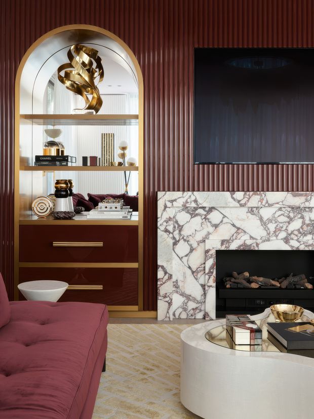A luxurious living room featuring a deep burgundy vertical fluted wall, a large black television, and a dramatic pale marble fireplace surround with dark veining. An arched built-in display cabinet with brass trim, glass shelving, decorative objects, books, and lower burgundy cabinetry stands beside the fireplace. The room includes a plush burgundy sofa, a rounded white coffee table, a patterned neutral rug, and polished brass accents. The overall composition is refined, glamorous, and richly layered.