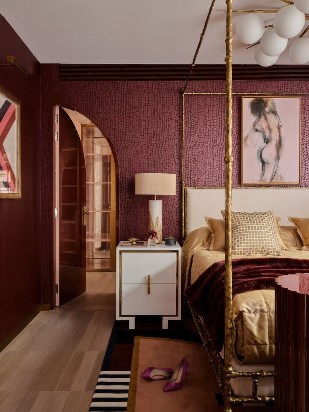 A richly styled bedroom with deep burgundy textured wallcovering, a brass four-poster bed, layered cream and burgundy bedding, and a white bedside cabinet with brass detailing. An arched doorway opens to a timber-lined adjoining area, while framed artwork, a sculptural globe pendant, and a striped area rug add decorative character. The room has a dramatic, luxurious feel with warm lighting and refined vintage-inspired detailing.