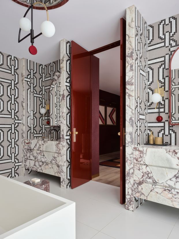 A dramatic contemporary bathroom featuring full-height black-and-white geometric mosaic tile walls, richly veined marble vanity surfaces, and a white freestanding bathtub in the foreground. Deep red glossy doors, wall panels, and trim contrast with the monochrome tilework, while sculptural globe pendant lights add an art-deco-inspired decorative element. A darker adjoining room is visible through the open doorway.