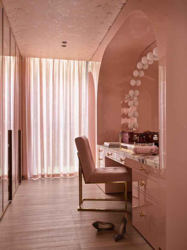 A glamorous blush-pink dressing and vanity area with a glossy built-in desk, drawers, arched mirror, gold hardware, and a tufted upholstered chair. Floor-to-ceiling sheer curtains bring soft daylight into the narrow room, while decorative round wall lights and a veined marble-like ceiling add an opulent, highly coordinated finish.
